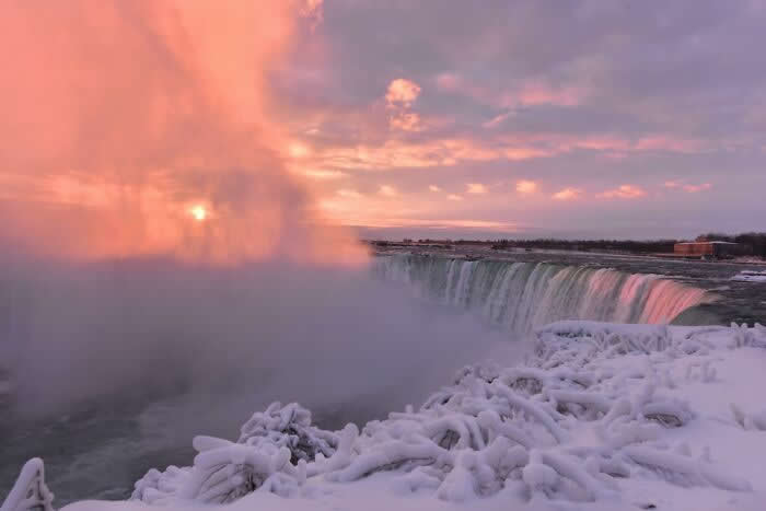 Soft pink sunrise light spreads across Niagara Falls as icy mist rises above snow-covered formations and frozen branches during a peaceful winter morning.