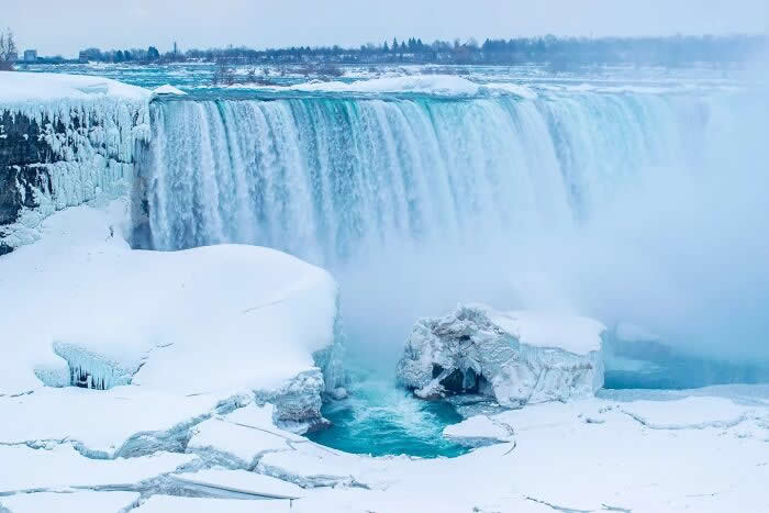 Frozen Niagara Falls cascades into icy blue water surrounded by snow-covered rock formations and thick winter mist under a pale cloudy sky.