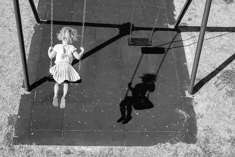 The Art of Perfect Timing: 31 Stunning Street Photos Taken at Exactly the Right Moment 44 Black-and-white photograph of a young girl on a swing viewed from above. Her shadow appears on the playground surface beside an empty swing, creating a striking visual relationship through light, timing, and composition.