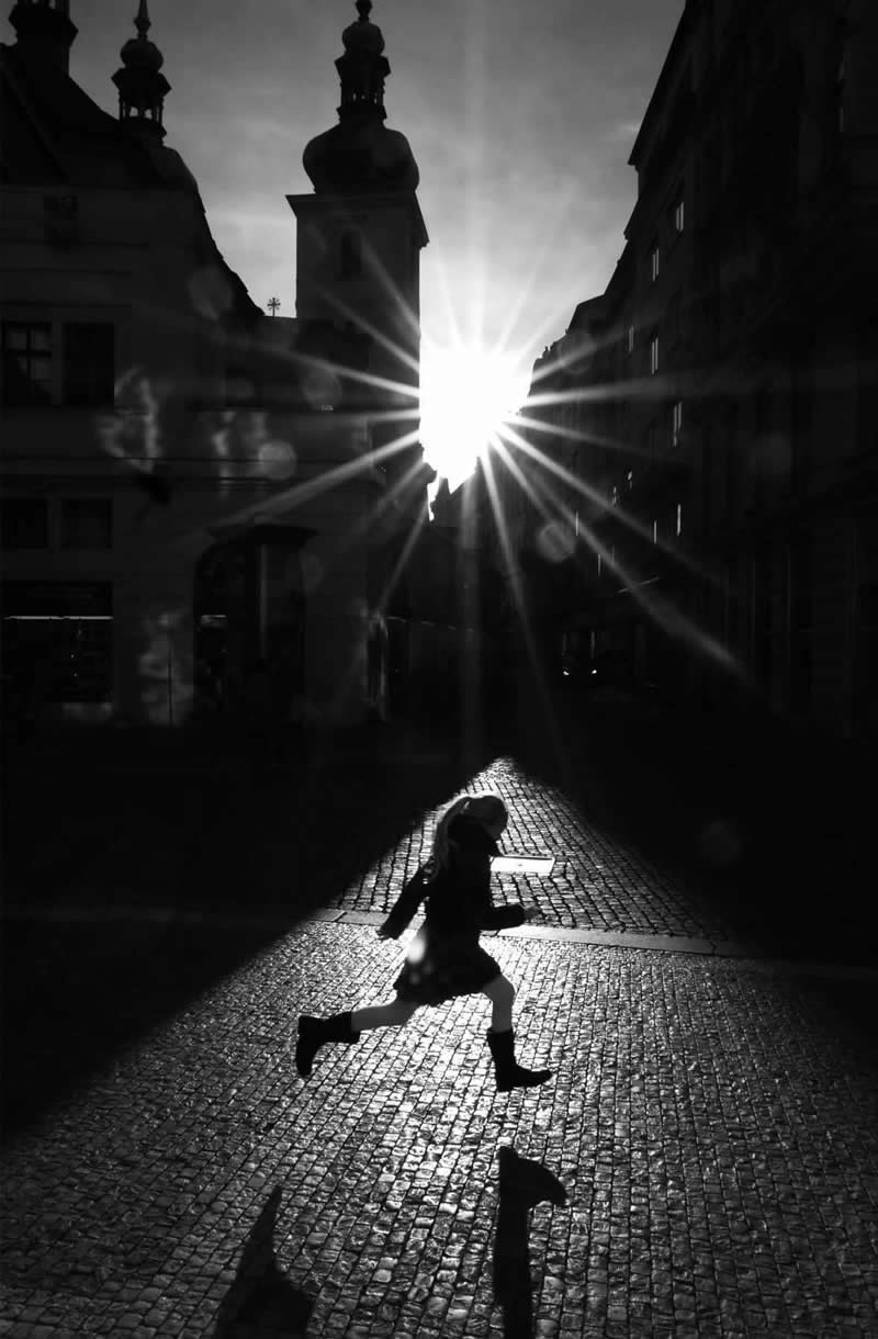 The Art of Perfect Timing: 31 Stunning Street Photos Taken at Exactly the Right Moment 45 Black-and-white street photograph of a person running across a cobblestone square as sunlight bursts between historic buildings. Strong silhouettes, starburst light rays, long shadows, and precise timing create a dramatic and visually striking urban scene.