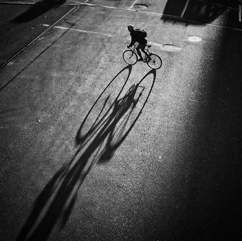 The Art of Perfect Timing: 31 Stunning Street Photos Taken at Exactly the Right Moment 47 Black-and-white street photograph of a cyclist riding across a sunlit road, casting a long elongated shadow. Viewed from above, the image emphasizes dramatic light, strong contrast, geometric shapes, and a perfectly timed urban moment.