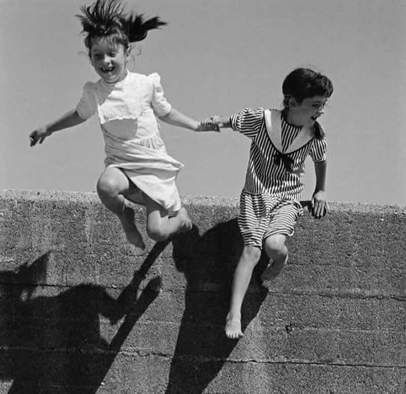The Art of Perfect Timing: 31 Stunning Street Photos Taken at Exactly the Right Moment 48 Black-and-white photograph of two young girls holding hands while jumping from a stone wall. One child is captured mid-air with hair flying upward, while strong shadows below add depth, movement, and dramatic visual impact.