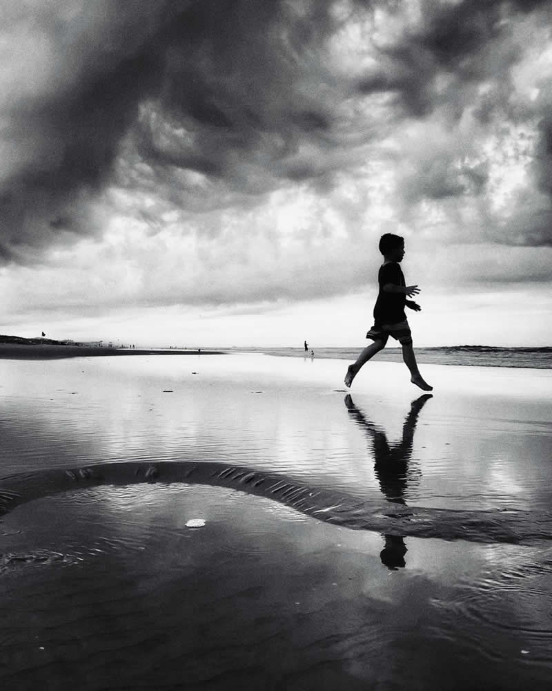 The Art of Perfect Timing: 31 Stunning Street Photos Taken at Exactly the Right Moment 49 Black-and-white photograph of a child running across a wet beach beneath dramatic storm clouds. Reflections shimmer on the water-covered sand, while the child’s silhouette and mirrored image create a dynamic composition filled with movement and atmosphere.
