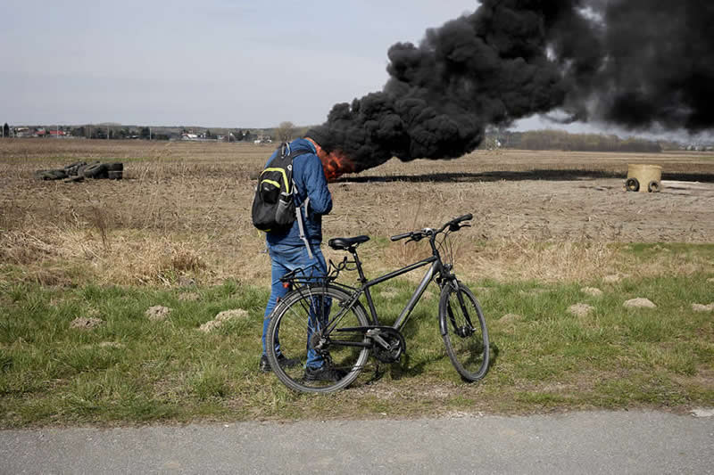 The Art of Perfect Timing: 31 Stunning Street Photos Taken at Exactly the Right Moment 51 Color street photograph of a man standing beside a bicycle in a rural field while thick black smoke from a distant fire aligns behind his head. Precise timing and composition create a striking visual illusion against the open landscape.