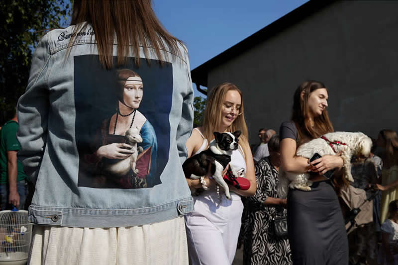 The Art of Perfect Timing: 31 Stunning Street Photos Taken at Exactly the Right Moment 52 Color street photograph showing a woman wearing a denim jacket printed with a classical portrait holding an animal, while nearby women hold small dogs. Clever timing and framing create a visual connection between the artwork and real-life scene.