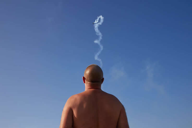 The Art of Perfect Timing: 31 Stunning Street Photos Taken at Exactly the Right Moment 53 Minimalist photograph of a bald man viewed from behind against a clear blue sky, with a winding smoke trail from an aircraft perfectly aligned above his head, creating the illusion of a thought or plume rising upward.