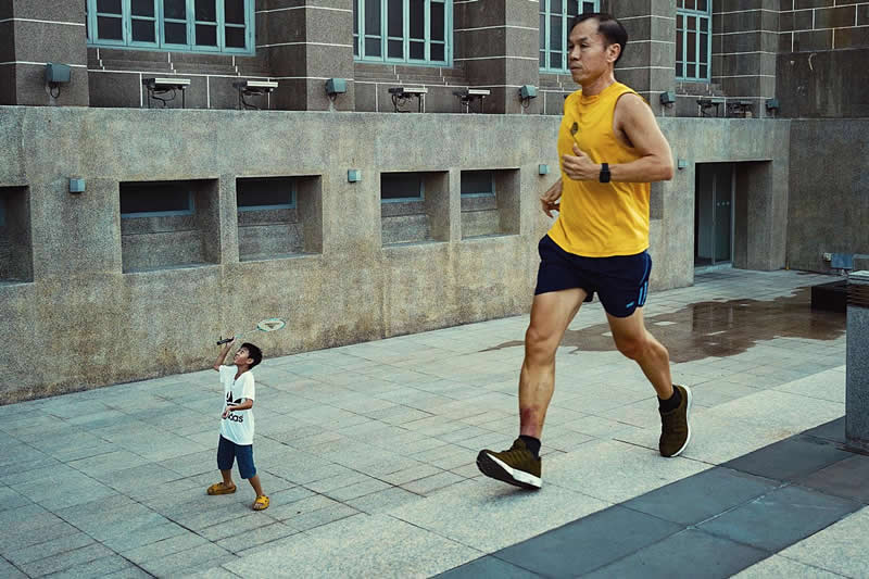 The Art of Perfect Timing: 31 Stunning Street Photos Taken at Exactly the Right Moment 54 Street photograph showing a man jogging past a building while a young child stands nearby holding a toy airplane, captured from a perspective that makes the runner appear giant-sized compared to the child.