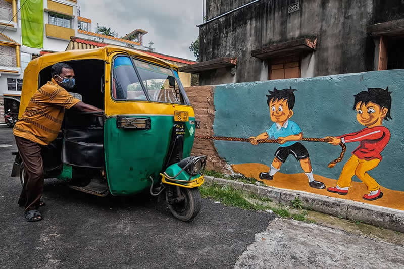 The Art of Perfect Timing: 31 Stunning Street Photos Taken at Exactly the Right Moment 55 Street photograph of an auto-rickshaw parked beside a colorful wall mural of two children pulling a rope, captured from an angle that makes it appear as though the painted children are pulling the vehicle.