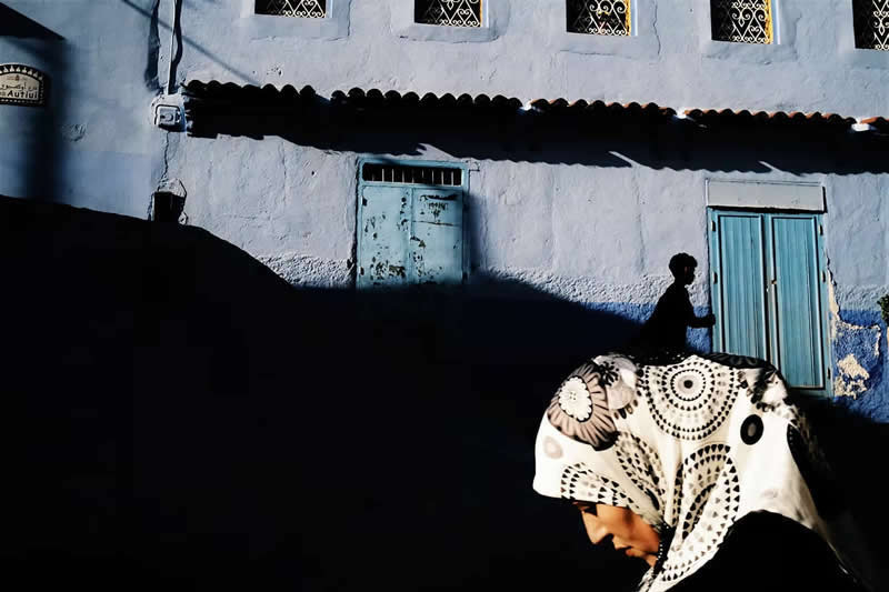 The Art of Perfect Timing: 31 Stunning Street Photos Taken at Exactly the Right Moment 56 Street photograph featuring a woman in a patterned headscarf walking through dramatic shadows, with a silhouetted figure in the background and blue-painted building walls creating strong contrast and layered composition.
