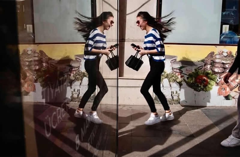 The Art of Perfect Timing: 31 Stunning Street Photos Taken at Exactly the Right Moment 57 Street photograph of a woman walking beside a reflective glass surface, creating a mirror image that makes it appear as though two identical women are facing each other in perfect symmetry.