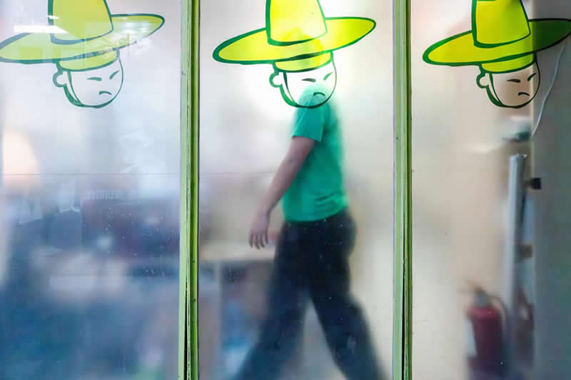 The Art of Perfect Timing: 31 Stunning Street Photos Taken at Exactly the Right Moment 59 Street photograph of a person walking behind frosted glass panels, perfectly aligned with a cartoon figure wearing a large green hat, creating the illusion that the illustrated hat belongs to the passerby.
