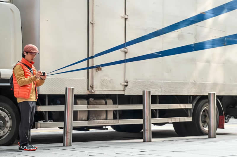 The Art of Perfect Timing: 31 Stunning Street Photos Taken at Exactly the Right Moment 60 Street photograph of a man holding a camera beside a parked truck, with blue stripes on the truck perfectly aligned to appear as though they extend directly from the camera lens.