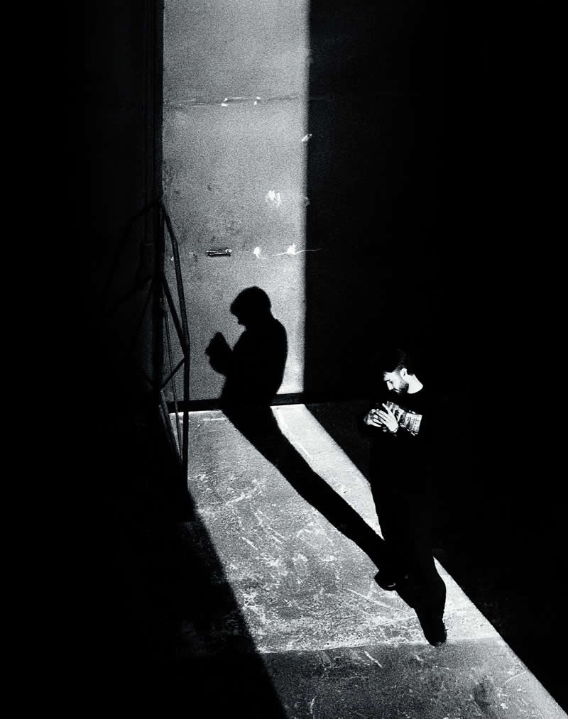 The Art of Perfect Timing: 31 Stunning Street Photos Taken at Exactly the Right Moment 38 Black-and-white street photograph of a person standing in a bright shaft of light surrounded by deep shadows. The subject’s elongated shadow stretches across the illuminated floor, creating dramatic contrast, strong geometry, and a perfectly timed urban scene.