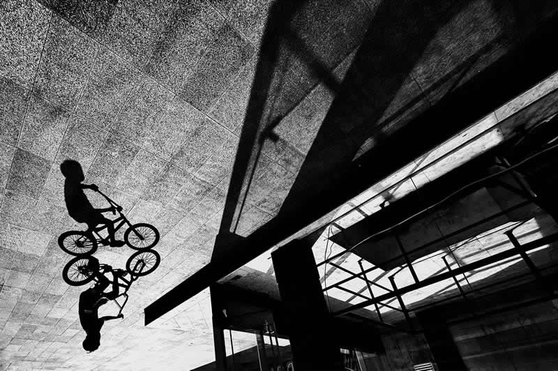 The Art of Perfect Timing: 31 Stunning Street Photos Taken at Exactly the Right Moment 43 Black-and-white street photograph of a cyclist and their reflection captured on a reflective surface beneath an urban structure. Dramatic shadows, geometric lines, and an unusual perspective create a striking composition emphasizing timing, symmetry, and urban design.