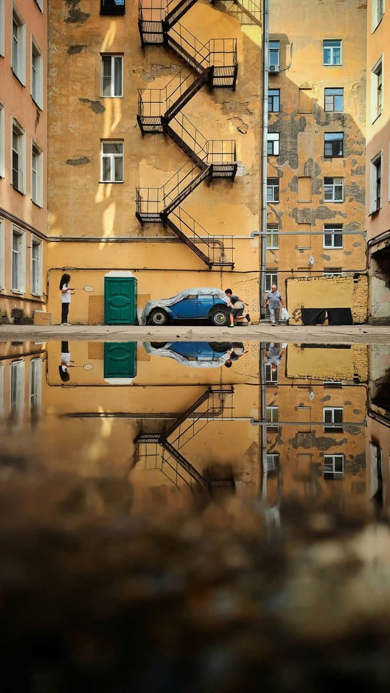The Mobile Revolution: What the Winners of 35AWARDS Tell Us About the Future of Phone Cameras 40 An urban courtyard surrounded by aging apartment buildings features a zigzag fire escape staircase, a parked blue car, and several people. A large puddle in the foreground reflects the entire scene, creating strong symmetry, depth, and a visually captivating composition.