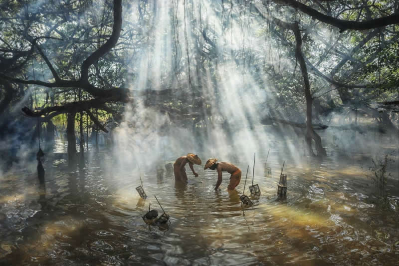 The Mobile Revolution: What the Winners of 35AWARDS Tell Us About the Future of Phone Cameras 31 Two people stand in shallow water beneath a dense forest canopy as dramatic rays of sunlight stream through the trees. Reflections ripple across the water, while mist and light create a serene, ethereal atmosphere rich in depth and beauty.