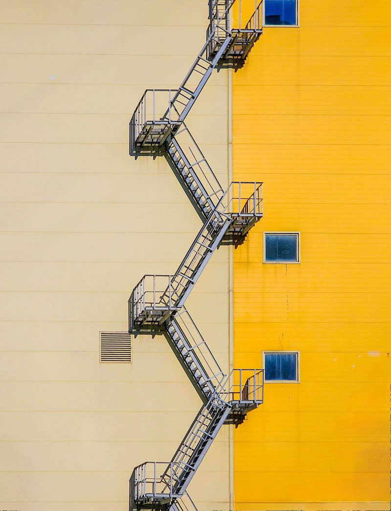 The Mobile Revolution: What the Winners of 35AWARDS Tell Us About the Future of Phone Cameras 49 A metal fire escape staircase zigzags vertically across the junction of two building walls, one cream-colored and the other bright yellow. Small windows and clean geometric lines create a minimalist architectural composition emphasizing symmetry, contrast, pattern, and visual balance.