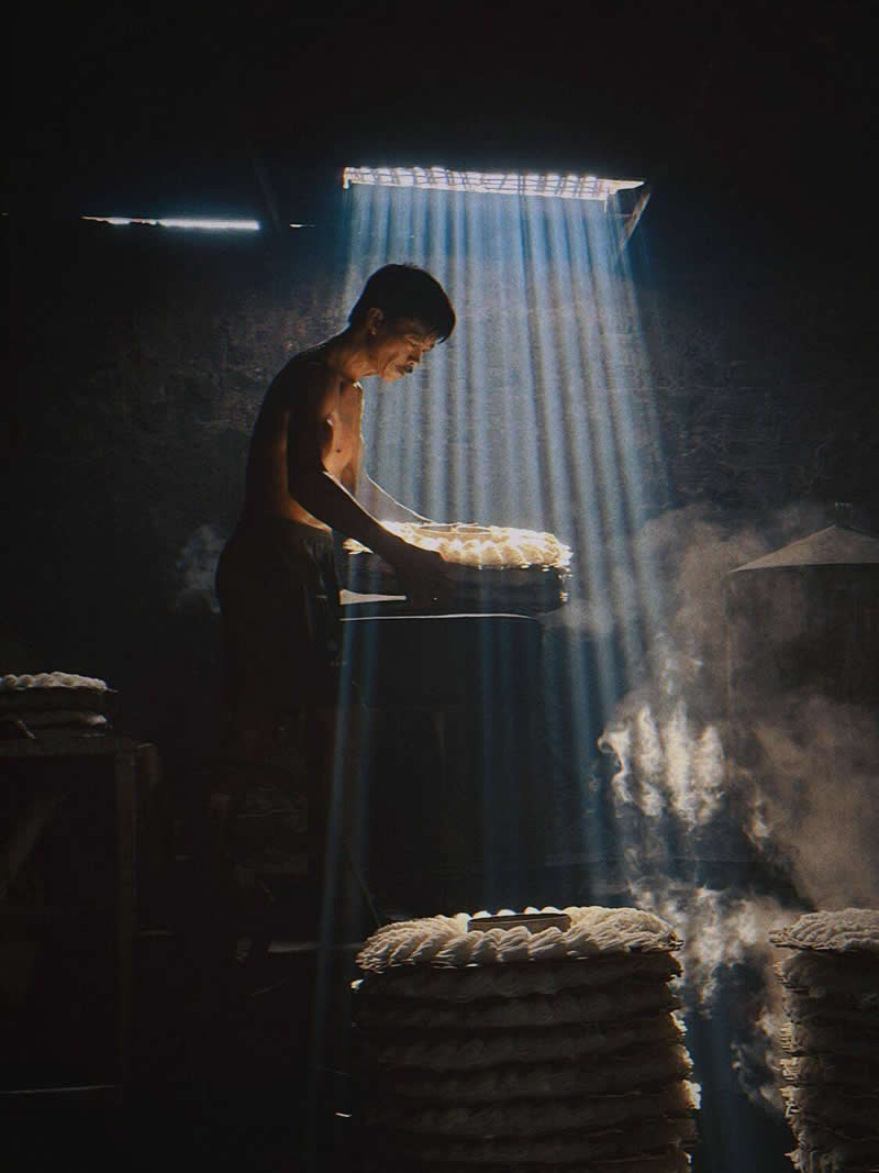 The Mobile Revolution: What the Winners of 35AWARDS Tell Us About the Future of Phone Cameras 32 A worker stands indoors beside steaming trays, illuminated by powerful rays of sunlight streaming through a roof opening. The dark surroundings contrast with the bright beams and drifting steam, creating a dramatic, atmospheric scene rich in texture and emotion.