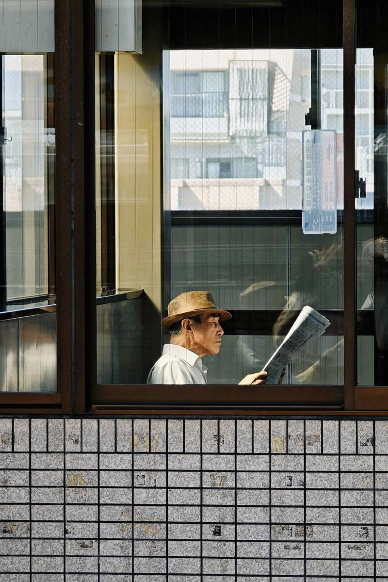 The Mobile Revolution: What the Winners of 35AWARDS Tell Us About the Future of Phone Cameras 38 A man wearing a hat sits indoors reading a newspaper, viewed through large window frames. Geometric lines, reflections on the glass, and warm natural light create a layered composition that highlights contemplation, urban life, and visual balance.