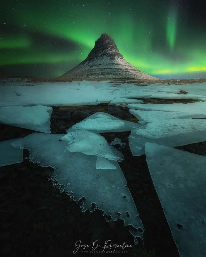 A snow-covered mountain rises beneath green aurora lights, with broken ice sheets filling the foreground and a starry night sky stretching across the scene.