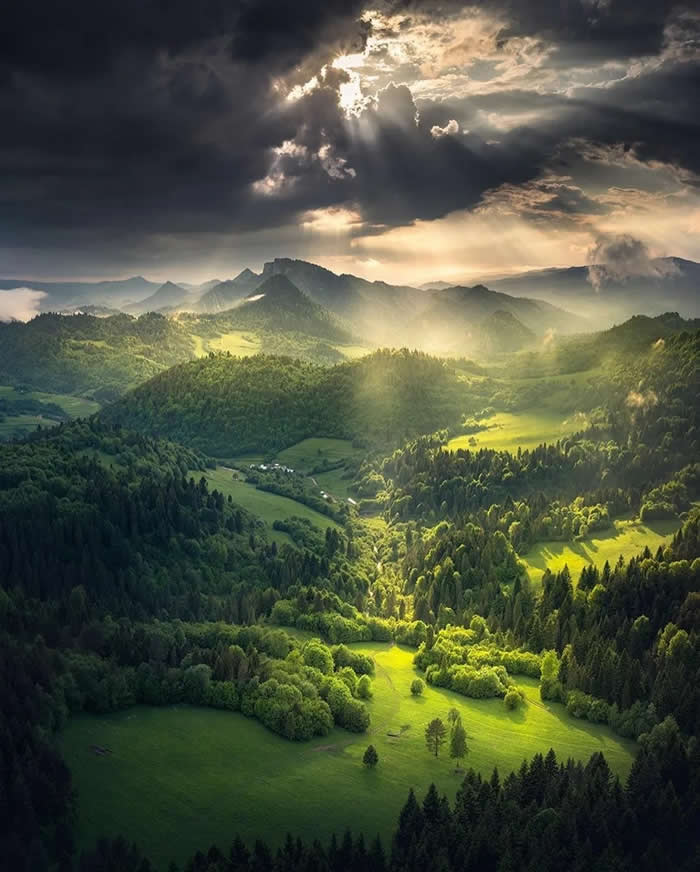 Sun rays stream through dark storm clouds onto a lush green valley filled with forests, rolling hills, and distant mountain ridges, creating bright patches of light across the landscape.