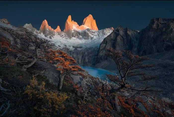 Snow-capped mountain peaks glow orange at sunset above a rocky landscape with twisted trees and a turquoise lake nestled in the valley below.