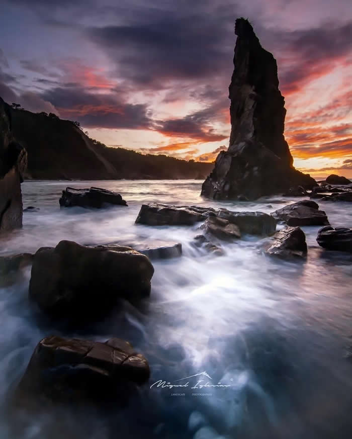 A tall sea stack rises from the ocean at sunset, surrounded by dark rocks and silky long-exposure water beneath a dramatic purple and orange sky.