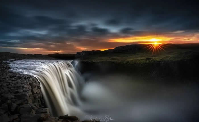 A wide waterfall pours over a rocky cliff at sunset, with silky mist below, dark clouds overhead, and a bright starburst sun glowing on the horizon.