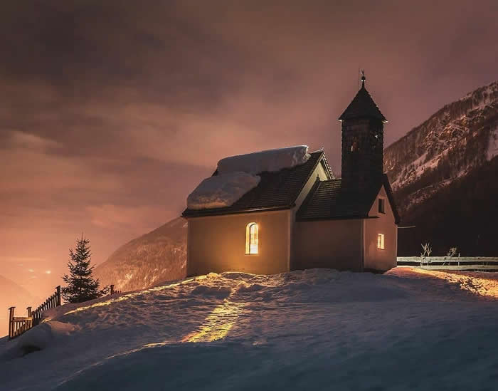 A small chapel with glowing windows stands on a snowy hillside at dusk, with warm light in the sky and dark mountain slopes rising behind it.
