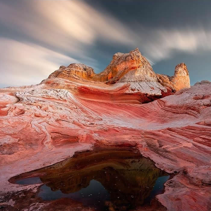 Layered red and white sandstone formations curve toward rocky peaks under a dramatic sky, with a dark reflective pool in the foreground adding depth to the desert landscape.
