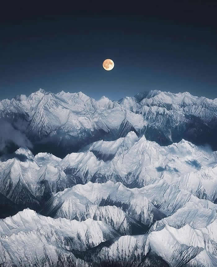 A full moon glows above a vast range of snow-covered mountain peaks under a deep blue night sky, with layered ridges stretching across the frame.