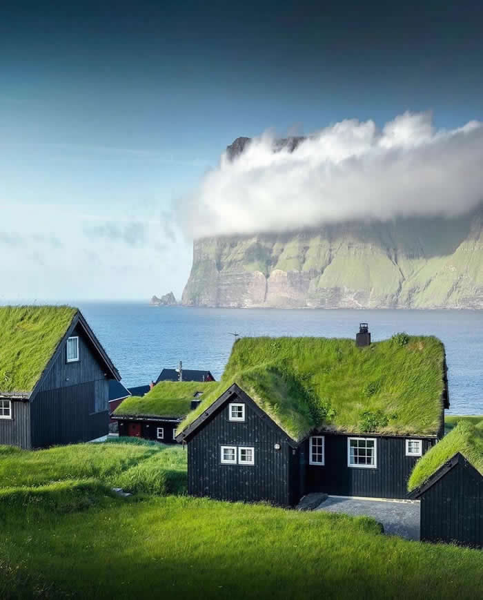 Black wooden houses with green grass roofs sit beside the sea, with a massive cliff partly covered by low clouds rising in the background under a clear blue sky.