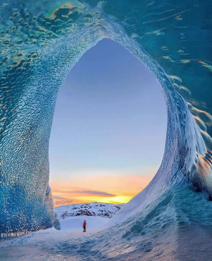 A small person stands on snowy ground beneath a massive blue ice arch, with the opening framing a glowing sunset sky and snow-covered landscape in the distance.