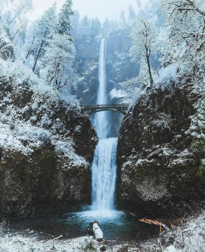 A tall waterfall plunges through a snowy forested gorge, passing beneath a stone bridge before dropping into a pool, with frost-covered trees and dark cliffs surrounding the scene.