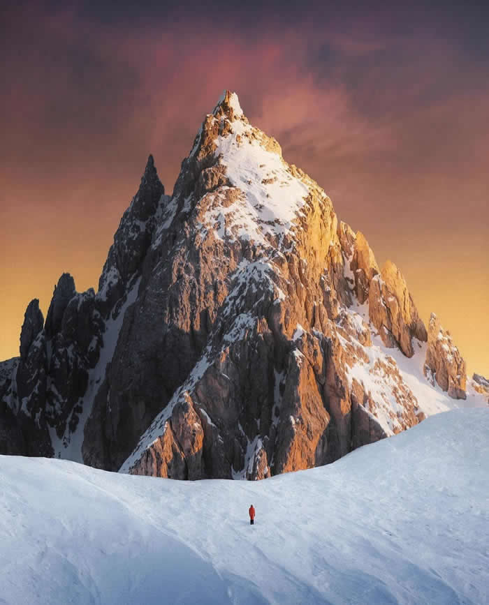 A tiny person in a red jacket stands on a snowy slope beneath a towering rocky mountain peak lit by warm sunset light, with a glowing pink-orange sky in the background.