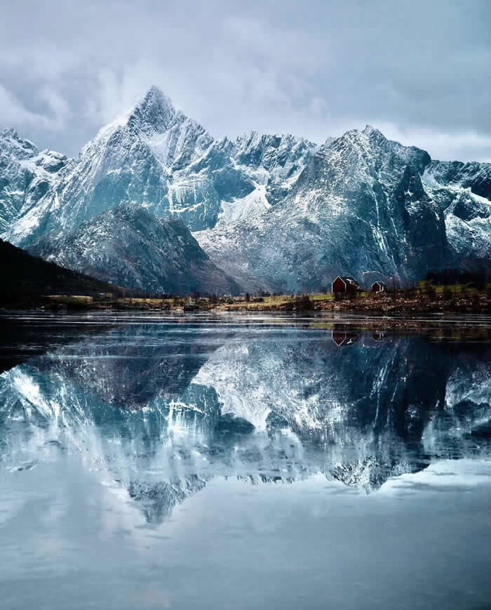 Snow-covered mountains rise above a calm fjord, reflected almost perfectly in the water, with a few small red cabins along the shoreline beneath the towering peaks.