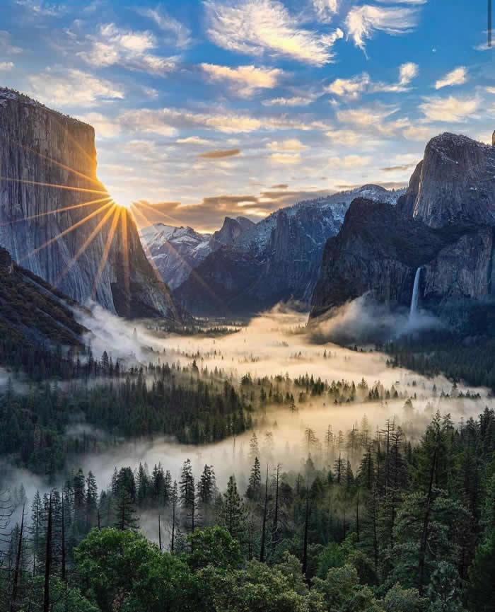 Sunrise lights Yosemite Valley with a starburst beside a towering granite cliff, while mist drifts above the forest below and a waterfall cascades down the opposite rock face.