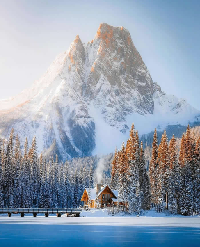 A small wooden cabin sits beside a frozen blue lake and snow-covered pine forest, with a massive sunlit mountain rising dramatically behind under a clear winter sky.