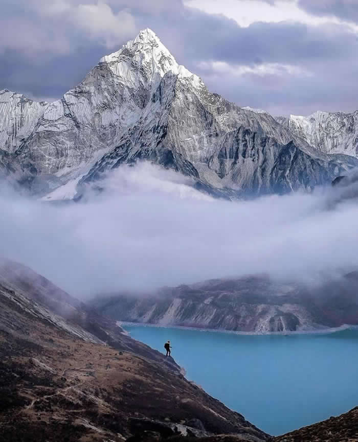 A lone hiker stands on a rocky slope above a bright turquoise lake, with low clouds drifting beneath a towering snow-covered mountain under a soft blue and lavender sky.