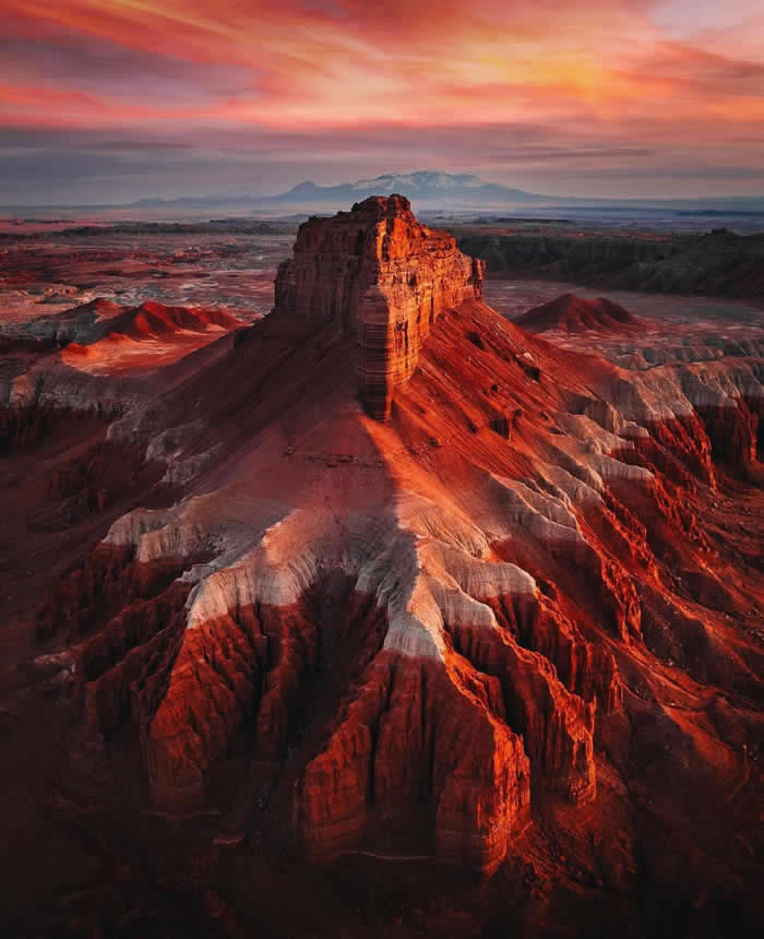 A towering red desert butte glows in warm sunset light, surrounded by deeply eroded canyons and layered rock formations beneath a dramatic pink and orange sky.
