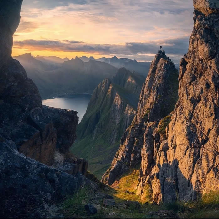 A dramatic mountain landscape at sunset seen between rocky cliffs, with a lone person standing on a sharp peak above a fjord and distant mountain ridges.