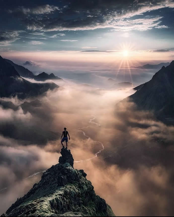 A person stands on a narrow rocky peak above a cloud-filled mountain valley at sunrise, with soft golden light, distant ridges, and a winding river visible through the mist.