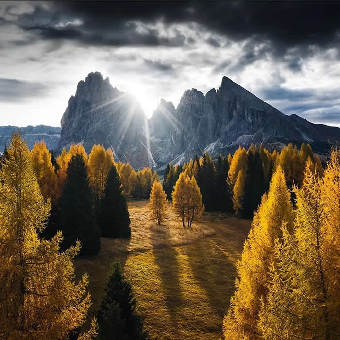Golden autumn trees stand in a sunlit valley below dramatic dark mountain peaks, with sunlight breaking through the rocks under a moody cloudy sky.