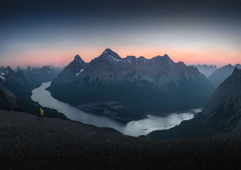 Blue Nights and Snowy Peaks: Cath Simard's 25 Dreamlike Landscapes of the Canadian Rockies 38 A lone hiker in a yellow jacket walks across a rocky ridge overlooking winding lakes and towering mountain peaks at dawn in the Canadian Rockies beneath a soft pastel sky.