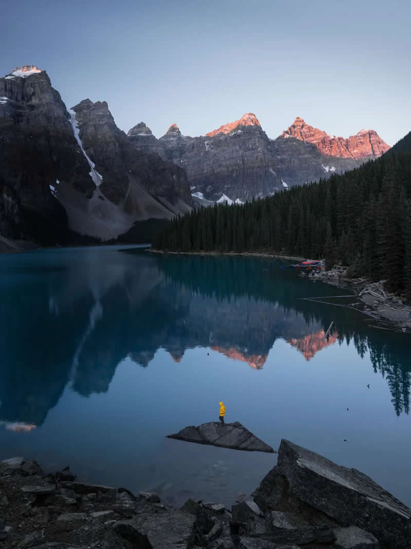 Blue Nights and Snowy Peaks: Cath Simard's 25 Dreamlike Landscapes of the Canadian Rockies 39 A person in a yellow jacket stands on a rock in a calm turquoise lake, reflecting sunlit mountain peaks, evergreen forests, and the serene beauty of the Canadian Rockies at sunrise.