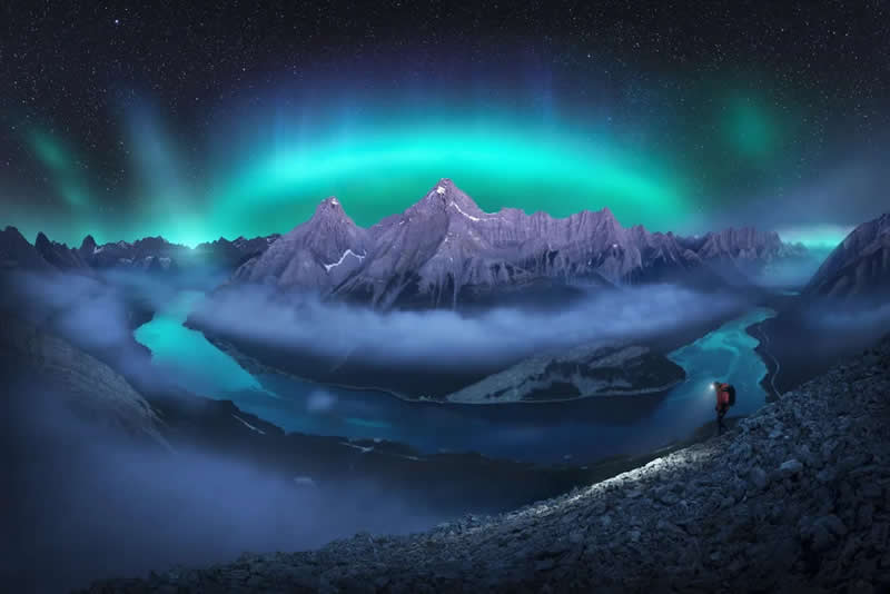 Blue Nights and Snowy Peaks: Cath Simard's 25 Dreamlike Landscapes of the Canadian Rockies 40 A lone hiker stands on a rocky ridge beneath the northern lights, overlooking winding rivers, mist-covered valleys, and dramatic mountain peaks in the Canadian Rockies under a star-filled night sky.
