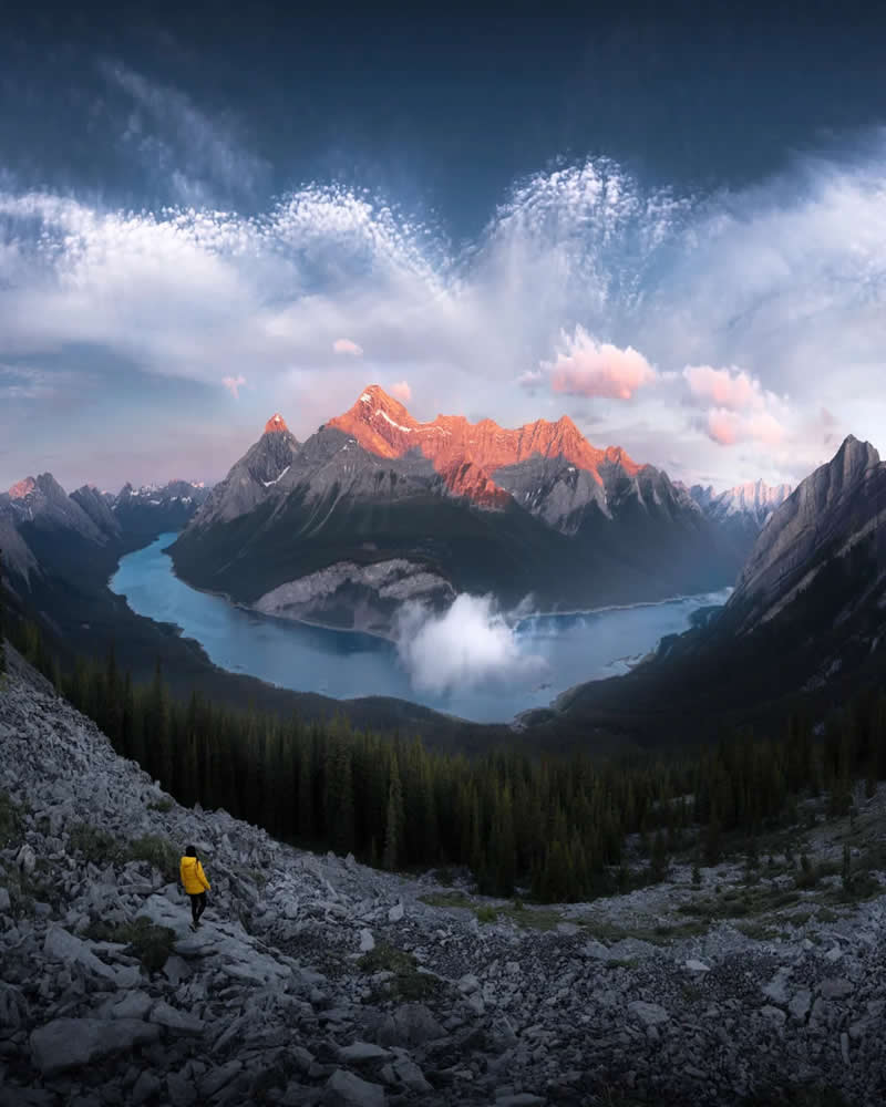 Blue Nights and Snowy Peaks: Cath Simard's 25 Dreamlike Landscapes of the Canadian Rockies 41 A lone hiker in a yellow jacket walks down a rocky mountainside overlooking winding turquoise lakes and sunrise-lit peaks beneath dramatic clouds in the Canadian Rockies.