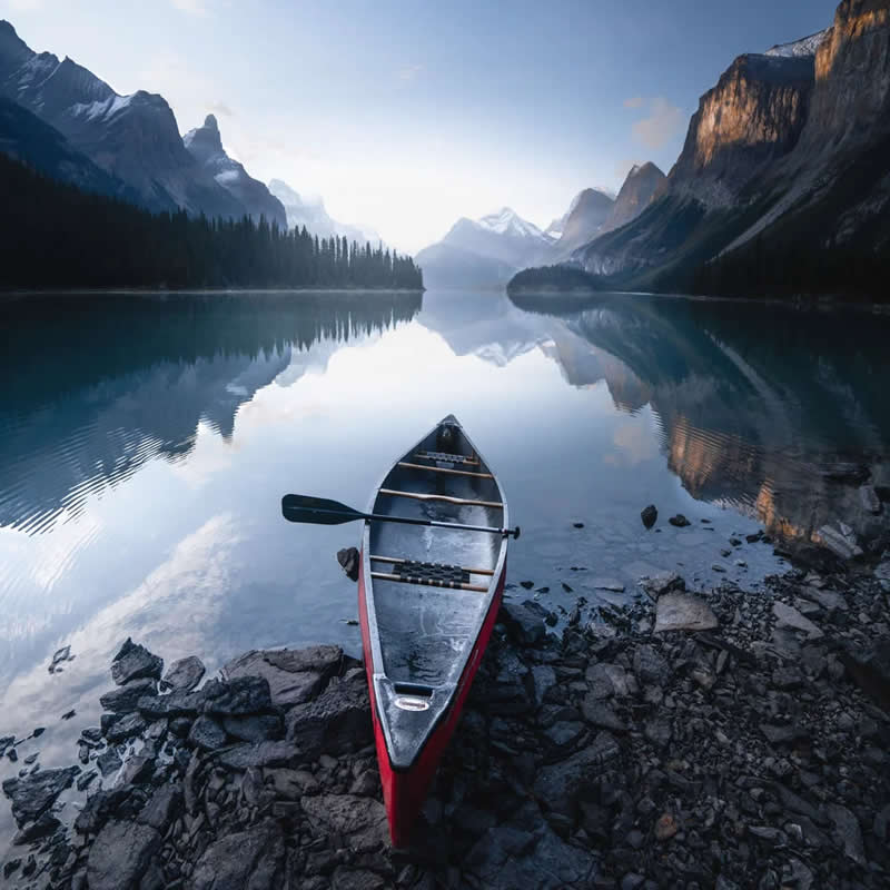 Blue Nights and Snowy Peaks: Cath Simard's 25 Dreamlike Landscapes of the Canadian Rockies 42 A red canoe rests on the rocky shore of a calm alpine lake, reflecting towering mountains and evergreen forests beneath a clear morning sky in the Canadian Rockies.