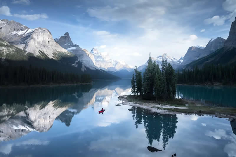 Blue Nights and Snowy Peaks: Cath Simard's 25 Dreamlike Landscapes of the Canadian Rockies 43 A lone red canoe floats beside a small tree-covered island on a calm alpine lake, reflecting snow-capped mountains, evergreen forests, and a peaceful sky in the Canadian Rockies.