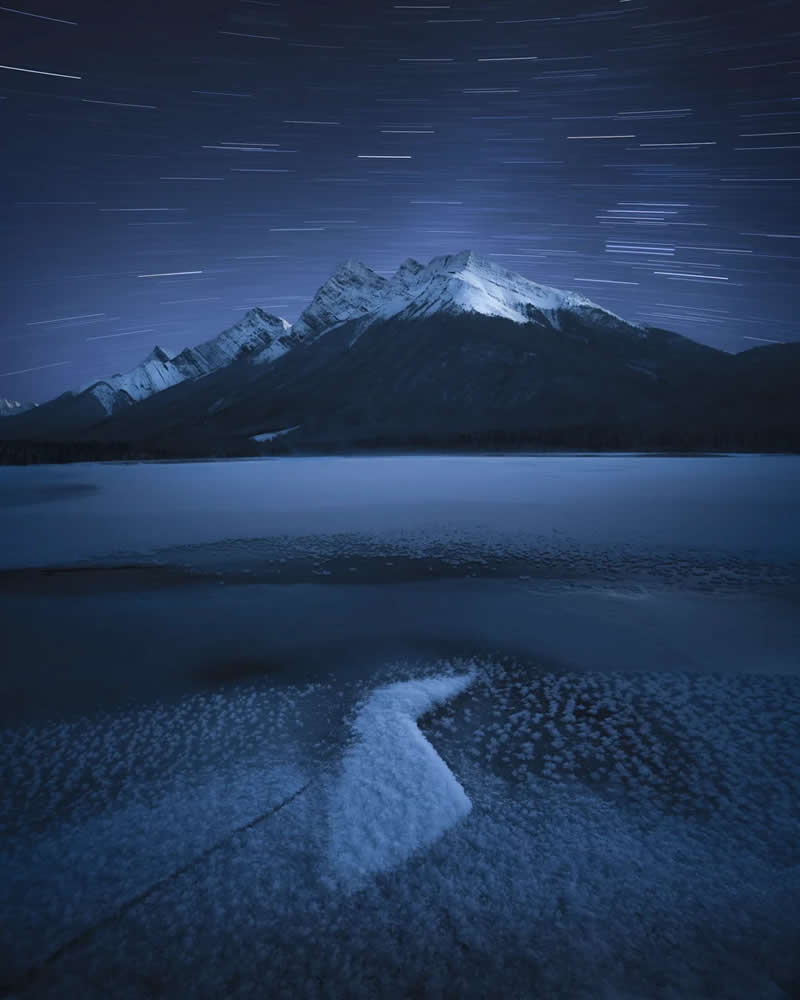 Blue Nights and Snowy Peaks: Cath Simard's 25 Dreamlike Landscapes of the Canadian Rockies 44 Star trails arc across the night sky above snow-covered mountain peaks and a frozen alpine lake, with textured ice in the foreground creating a serene Canadian Rockies winter landscape.